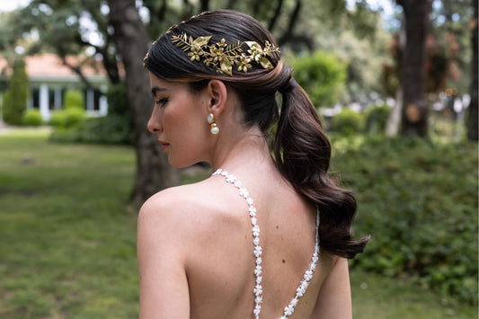 chica con melena larga y castaña recogida en una coleta con ondas. Lleva un vestido blanco de novia de tirantes con detalles, un cubre rayas dorado color oro con detalles verdes y unos pendientes ACUS de piedras y perlas naturales, latón bañado en oro.