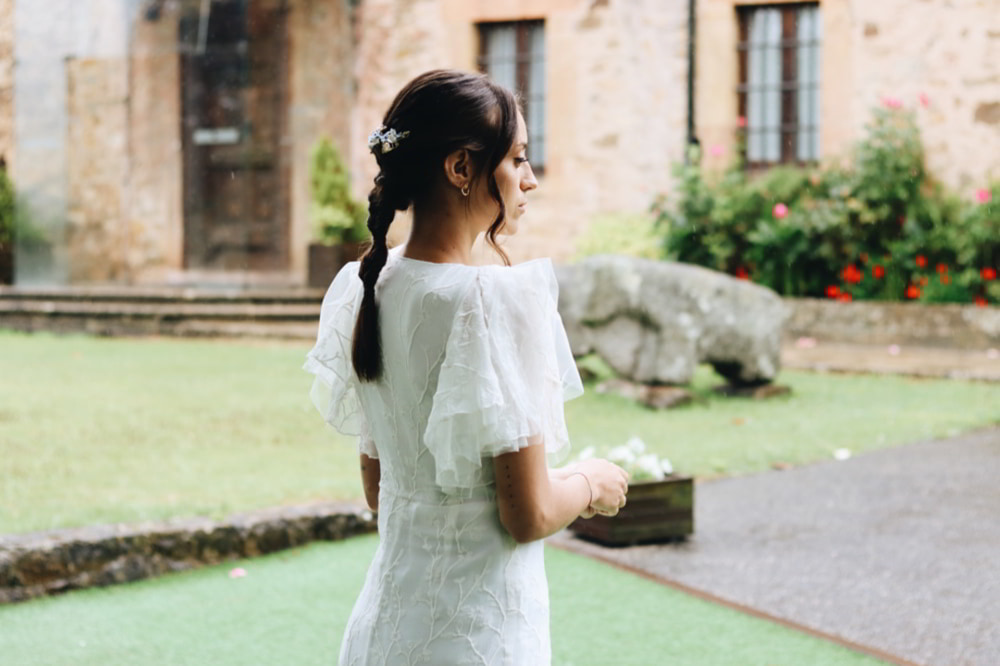 Una chica morena vestida de novia con un vestido blanco con mangas vaporosas. Lleva un recogido trenzado con un tocado en tonos blancos y unos pendientes ACUS pequeños.